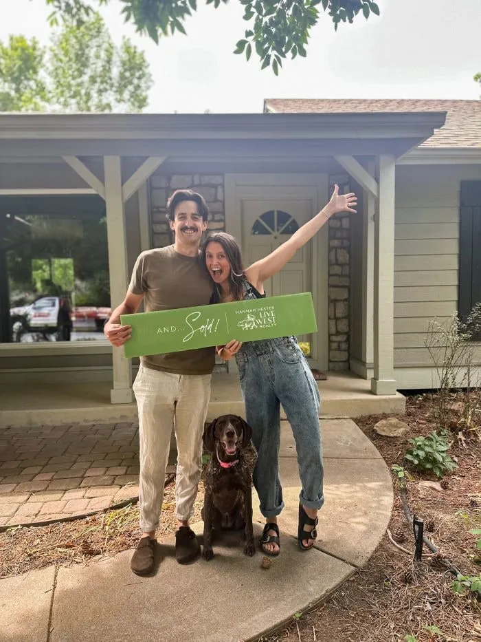 Two new homeowners and their dog celebrating with a Sold sign outside the house