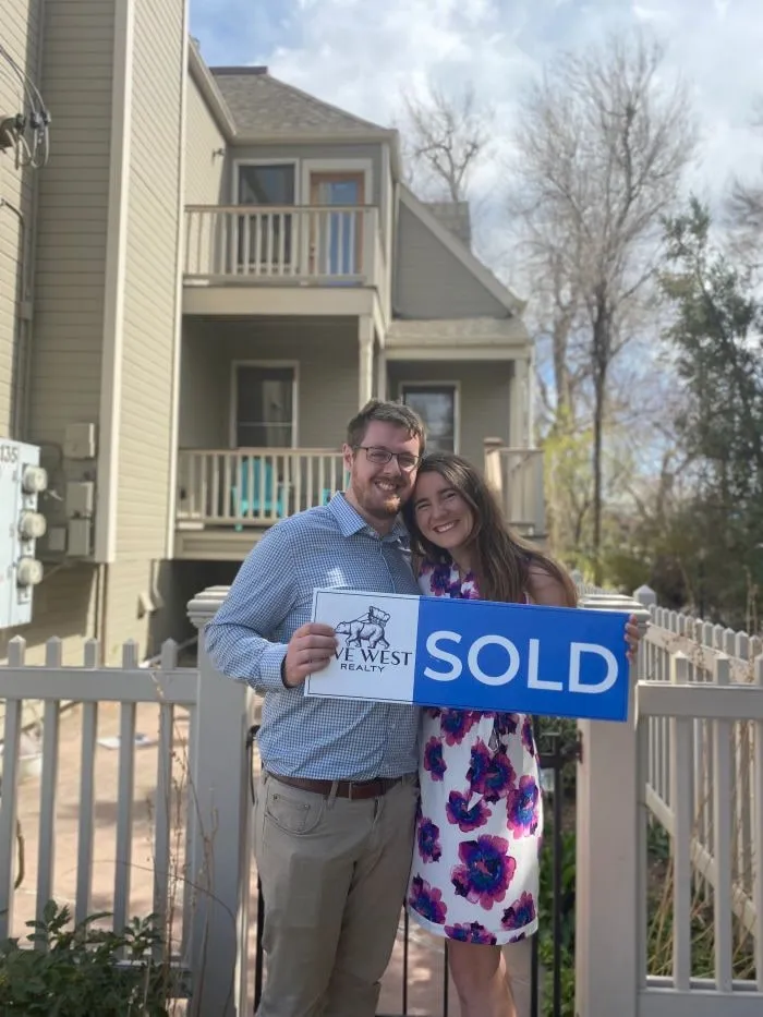 A couple holding a Live West SOLD sign by a white picket fence